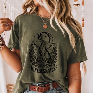 以下が含まれることがあります： オリーブグリーンの半袖Tシャツ。三日月の下に座るウサギ、その周りを囲む植物と松ぼっくりの黒いグラフィックプリントが施されており、下部には「WINTER SOLSTICE」という文字が太字のアーチ状フォントでプリントされています。