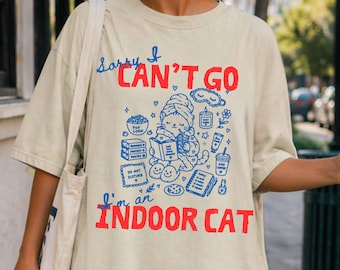 May include: A beige t-shirt featuring a graphic print with the text "Sorry I CAN'T GO I'm an INDOOR CAT" in red. The blue illustration depicts a cat wearing a towel on its head, reading a book, surrounded by icons of popcorn, a candle, and coffee.