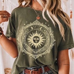 May include: An olive green short-sleeve t-shirt featuring a large, circular graphic print in a light yellow hue. The design depicts a central sun face surrounded by intricate floral patterns, crescent moons, and stars within a decorative border.