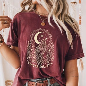Puede incluir: Una camiseta de manga corta color granate con un gráfico en color crema de un cuervo posado en una corona de follaje, una luna creciente y estrellas. El texto "WINTER SOLSTICE" está impreso en una fuente curva en la base del diseño.