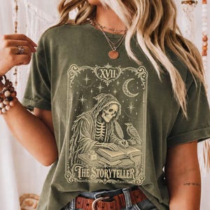 May include: An olive green t-shirt featuring a cream-coloured graphic of a skeleton in a hooded robe writing in a book. The design includes a raven, a crescent moon, stars, the Roman numeral XVII, and the text "THE STORYTELLER" in a frame.