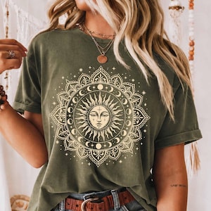 Könnte beinhalten: Ein olivgrünes Kurzarm-T-Shirt mit einem großen, cremefarbenen Grafikdruck eines Sonnengesichts, das von komplizierten Mandala-Mustern und Mondphasen umgeben ist. Das Design ist mittig auf der Brust platziert und wirkt sehr detailliert.