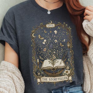May include: Dark grey t-shirt with a detailed illustration. The design includes an open book, butterflies, flowers, a lantern, and a banner with the text "THE STORYTELLER." The artwork is framed by a decorative border with the Roman numeral XXVII.