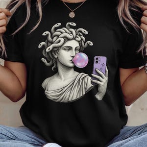 May include: Black t-shirt with a graphic of Medusa blowing a bubblegum bubble and taking a selfie with a purple phone. The Medusa illustration is in a classical statue style.