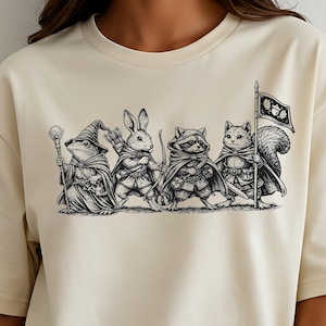 May include: Cream-colored t-shirt featuring a black and white illustration of four anthropomorphic animals: a frog, rabbit, raccoon, and squirrel. Each animal is dressed in fantasy attire, with the squirrel holding a flag.