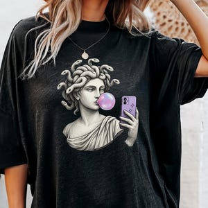 May include: Black oversized t-shirt with a graphic of a Medusa bust blowing a bubblegum bubble while taking a selfie with a purple phone. The Medusa bust is in a classical style with snakes for hair.
