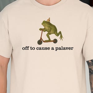 Könnte beinhalten: Beiges T-Shirt mit einer grünen Frosch-Illustration, der einen Partyhut trägt und Roller fährt. Unter der Grafik steht der Text "off to cause a palaver". Ein lässiges Unisex-T-Shirt.