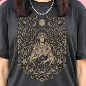 May include: Dark grey t-shirt with a detailed illustration of a woman holding a key, surrounded by floral and celestial elements. The design is in shades of brown and gold, with a full moon and crescent moons. The shirt has short sleeves and a crew neck.