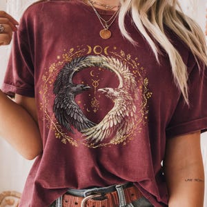 May include: Burgundy-colored t-shirt featuring a circular design with two stylized birds, one black and one white, facing each other. The design includes crescent moons and floral elements in gold. The shirt has rolled-up sleeves and a brown belt.