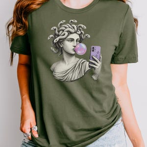 May include: Olive green t-shirt with a graphic of a Medusa bust blowing a bubblegum bubble while holding a purple smartphone. The Medusa bust is in a classical style, with snakes for hair. The bubble is pink.