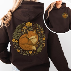 May include: A dark brown hoodie featuring a circular design on the back with a sleeping fox surrounded by yellow flowers, green leaves, and mushrooms. A smaller version of the design is on the front. The hoodie has a hood and a front pocket.