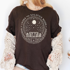 May include: A dark brown t-shirt with the text "UNDER THE SOLSTICE MOON" encircling a graphic of Stonehenge, a moon, stars, and celestial elements. The design is in a cream color. The shirt has rolled-up sleeves.