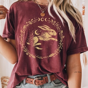 May include: Burgundy-colored t-shirt featuring a golden rabbit design within a circular floral wreath, moon phases, and stars. The shirt has short sleeves and a relaxed fit. A brown leather belt with a circular buckle is visible.