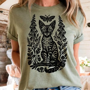 Könnte beinhalten: Salbeigrünes T-Shirt mit einem schwarzen Grafikdesign einer Katze, eines Schmetterlings und floraler Elemente. Die Katze ist zentral, mit einem Schmetterling darüber und Blumen auf beiden Seiten. Das Design hat eine Vintage- und botanische Ästhetik.