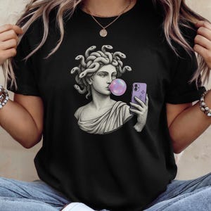 May include: Black t-shirt with a graphic of a Medusa bust blowing a bubblegum bubble while holding a purple smartphone. The Medusa bust is in shades of grey and white, with snakes for hair. The bubble is pink and purple.