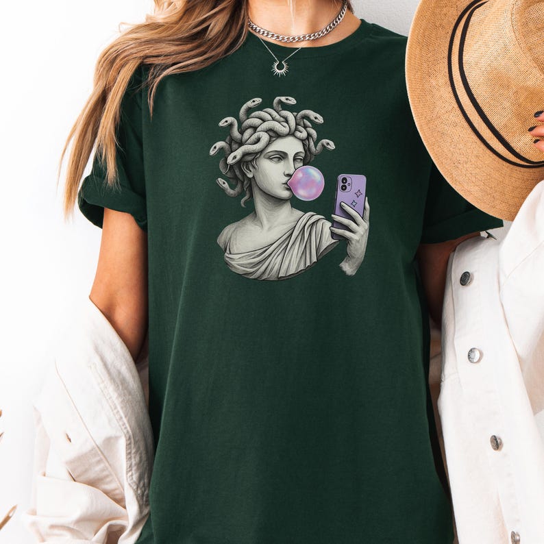 May include: Dark green t-shirt with a graphic of Medusa blowing a bubblegum bubble while taking a selfie with a purple phone. The Medusa graphic is in shades of grey and white.