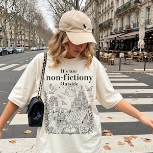 Könnte beinhalten: Eine beige Baseballkappe mit kleinem dunklem Logo und ein übergroßes cremefarbenes T-Shirt mit schwarzem Grafikdruck eines Schlosses, eines Paares und eines offenen Buches. Der Text lautet It's too non-fictiony Outside. Eine schwarze gesteppte Umhängetasche.