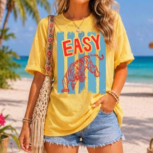May include: A yellow short-sleeved t-shirt featuring the word EASY in red capital letters above a graphic of a tiger. The design is set against vertical light blue stripes. The shirt has a crew neckline and a relaxed fit with rolled sleeves.