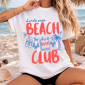 May include: White t-shirt with the words "Sandy pages BEACH CLUB" in red and blue. The design includes illustrations of flip-flops, a bikini, a book, and a palm tree. The shirt is on a beach setting.