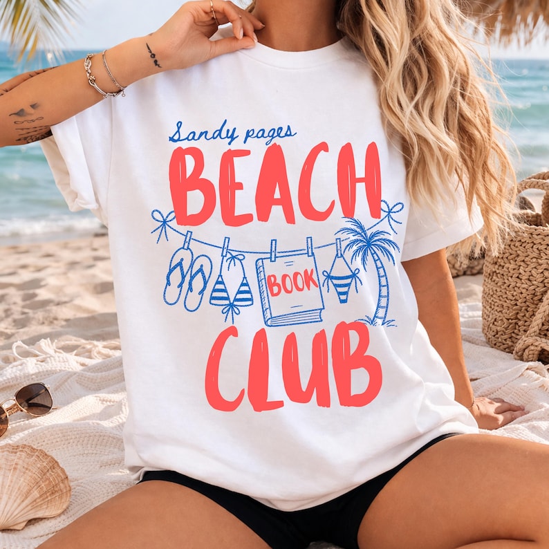 May include: White t-shirt with the words "Sandy pages BEACH CLUB" in red and blue. The design includes illustrations of flip-flops, a bikini, a book, and a palm tree. The shirt is on a beach setting.