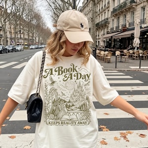 Może przedstawiać: Beżowa czapka z daszkiem z małym ciemnym logo oraz kremowy t-shirt oversize z ciemnozielonym nadrukiem graficznym przedstawiającym zamek, parę i otwartą książkę z napisem A BOOK A DAY KEEPS REALITY AWAY. Czarna pikowana torebka na ramię.