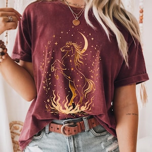 Op de afbeelding: Bordeauxrood t-shirt met een gouden illustratie van een steigerend paard, een halve maan en sterren. Het ontwerp bevat vlammen aan de voeten van het paard en een bladerenrand. Het shirt heeft korte mouwen en een ronde hals.