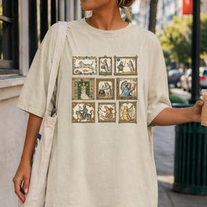 Op de afbeelding: Een beige t-shirt met korte mouwen voorzien van een raster van negen rechthoekige illustraties met katten in diverse middeleeuwse sc&egrave;nes, waaronder een kattenkoning, een kattenridder, een harp spelende kat en katten die koken en lezen.