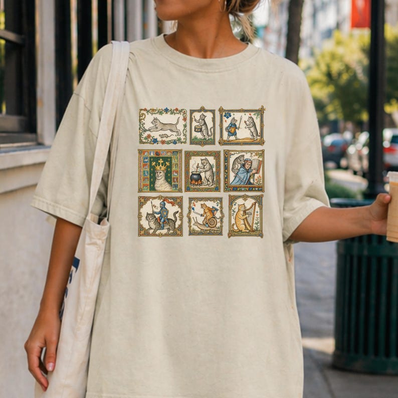 Op de afbeelding: Een beige t-shirt met korte mouwen voorzien van een raster van negen rechthoekige illustraties met katten in diverse middeleeuwse sc&egrave;nes, waaronder een kattenkoning, een kattenridder, een harp spelende kat en katten die koken en lezen.