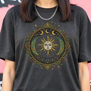 May include: Dark gray t-shirt featuring a celestial design. The graphic includes a sun with a face, crescent moons, and floral elements within a circular frame. The design is in shades of gold, green, and blue.