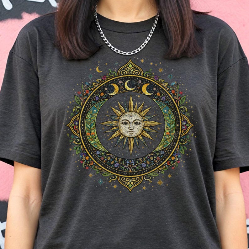 May include: Dark gray t-shirt featuring a celestial design. The graphic includes a sun with a face, crescent moons, and floral elements within a circular frame. The design is in shades of gold, green, and blue.