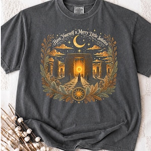 Może przedstawiać: Ciemnoszary t-shirt z nadrukiem przedstawiającym kamienny monument pod półksiężycem i rozgwieżdżonym niebem. Tekst "Have Yourself a Merry Little Solstice" jest wygięty w łuk nad sceną, która jest otoczona złotymi liśćmi i ikoną słońca.