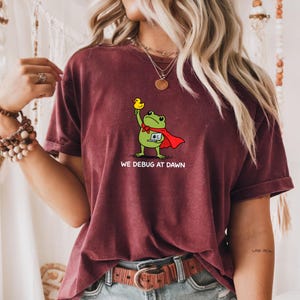 May include: A maroon short-sleeve t-shirt featuring a graphic of a green frog wearing a red cape, holding a yellow rubber duck, and using a laptop. Below the frog, the text reads WE DEBUG AT DAWN in white capital letters.