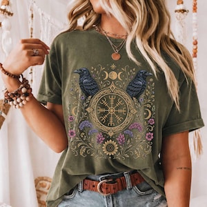 Könnte beinhalten: Ein olivgrünes Kurzarm-T-Shirt mit einem Grafikdruck von zwei schwarzen Raben, die ein zentrales, kreisförmiges goldenes Kompasssymbol flankieren. Das Design umfasst komplizierte Blumenranken mit violetten und rosa Blüten, Mondphasen und ein Sonnenmotiv.