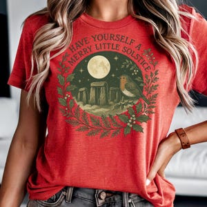 Könnte beinhalten: Rotes T-Shirt mit einem kreisförmigen Design mit Stonehenge, einem Vollmond, Sternen und einem Vogel. Der Text "Have Yourself a Merry Little Solstice" ist oben gewölbt. Das Design wird von einem Stechpalmenkranz umrahmt.