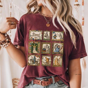 Op de afbeelding: Een bordeauxrood t-shirt met korte mouwen voorzien van een raster met negen rechthoekige illustraties van middeleeuws ogende katten die diverse activiteiten uitvoeren, zoals muziek maken, harnassen dragen en boeken lezen, in gouden kaders.