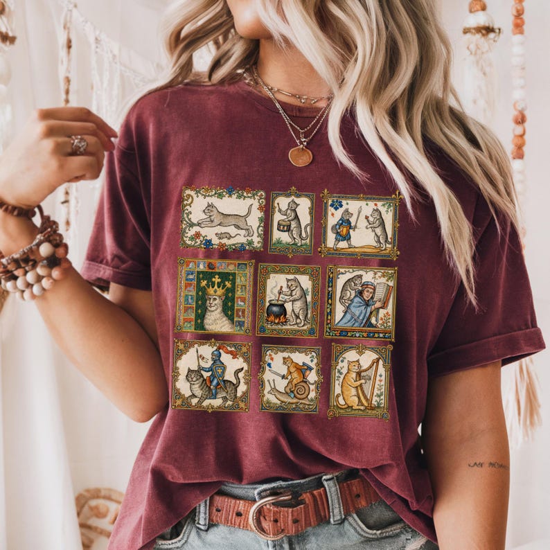 Op de afbeelding: Een bordeauxrood t-shirt met korte mouwen voorzien van een raster met negen rechthoekige illustraties van middeleeuws ogende katten die diverse activiteiten uitvoeren, zoals muziek maken, harnassen dragen en boeken lezen, in gouden kaders.