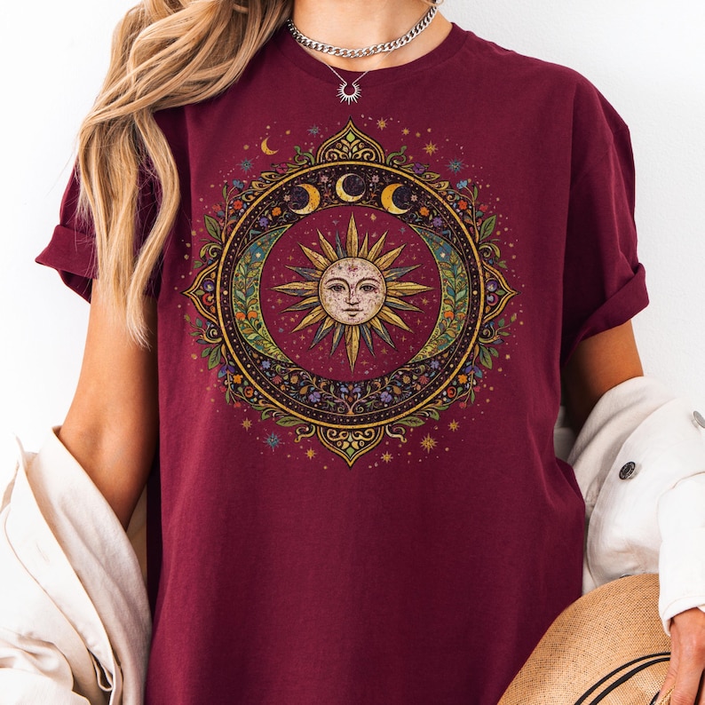 May include: Burgundy t-shirt featuring a detailed circular design with a sun face at the center, surrounded by crescent moons, stars, and floral elements. The design has a vintage, bohemian aesthetic.