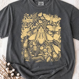 Può includere: Una t-shirt a maniche corte grigio antracite con una grande stampa grafica giallo chiaro di vari insetti, tra cui coleotteri, un ragno, una falena e un millepiedi, intervallati da elementi botanici come foglie, fiori e felci.