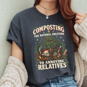 May include: A dark charcoal grey t-shirt featuring a graphic print of a Christmas tree growing out of a pile of dirt containing a skull wearing a Santa hat, bones, and mushrooms. The text reads: COMPOSTING THE NATURAL SOLUTION TO ANNOYING RELATIVES.