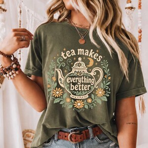 Puede incluir: Una camiseta de manga corta color verde oliva con un estampado gráfico de una tetera rodeada de flores y estrellas. El texto "tea makes everything better" está impreso con una fuente decorativa en el centro de la ilustración.