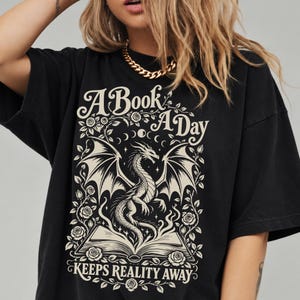 Könnte beinhalten: Ein schwarzes Kurzarm-T-Shirt mit einer cremefarbenen Grafik eines Drachen, der aus einem offenen Buch aufsteigt, umgeben von Rosen und Mondphasen. Der Text A BOOK A DAY KEEPS REALITY AWAY steht in einer dekorativen Schrift auf der Brust.