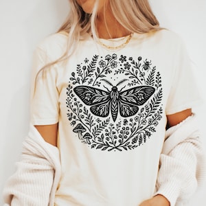 May include: Cream-colored t-shirt featuring a black moth design encircled by a wreath of flowers, leaves, and mushrooms. The moth has detailed wing patterns. The shirt has short sleeves and a crew neck.