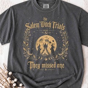 Può includere: Una t-shirt a maniche corte grigio antracite con una stampa grafica dorata. Il design include il testo "Salem Witch Trials 1692" e "They missed one" attorno a un'illustrazione centrale di una luna piena, un falò e due figure.