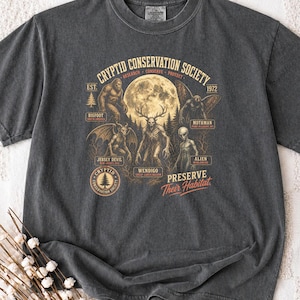 May include: A charcoal gray t-shirt featuring a graphic print titled Cryptid Conservation Society. The design includes illustrations of Bigfoot, Mothman, Jersey Devil, Wendigo, and an Alien, with the text Preserve Their Habitat and Est. 1972.