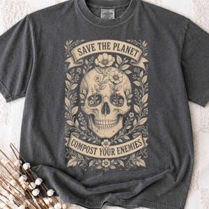 May include: A charcoal gray short-sleeve t-shirt featuring a cream-colored graphic of a human skull surrounded by floral vines. The text "SAVE THE PLANET" appears in a banner above the skull, and "COMPOST YOUR ENEMIES" is in a banner below.