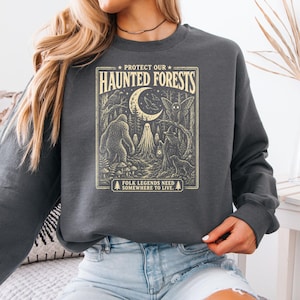 Puede incluir: Una sudadera gris oscuro de cuello redondo con un estampado gráfico color crema que incluye el texto "PROTECT OUR HAUNTED FORESTS" y "FOLK LEGENDS NEED SOMEWHERE TO LIVE." La ilustración muestra un sasquatch, un fantasma, una polilla y una figura.
