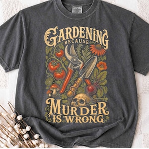May include: A charcoal grey t-shirt featuring a graphic print with the text "GARDENING BECAUSE MURDER IS WRONG". The design includes illustrations of pruning shears, a hand trowel, tomatoes, mushrooms, flowers, and a human skull in earthy tones.