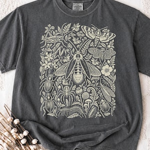 Op de afbeelding: Een antracietgrijs T-shirt met korte mouwen en een crèmekleurige grafische print van diverse insecten, waaronder een grote centrale kever, een mot, een spin en een duizendpoot, omringd door gedetailleerde botanische illustraties van bladeren en bloemen.