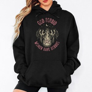 May include: A black hooded sweatshirt featuring a graphic print of four figures standing around a campfire with a pentagram symbol. The text "GOD FORBID WOMEN HAVE HOBBIES" is printed in a curved pink font above and below the central illustration.