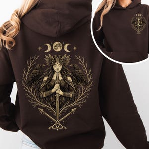 Puede incluir: Una sudadera con capucha marrón oscuro con una ilustración detallada en la espalda de una mujer sosteniendo una espada, flanqueada por cuervos, con un diseño de luna creciente y estrellas. La parte delantera tiene una versión más pequeña del diseño. La sudadera tiene capucha y bolsillo delantero.